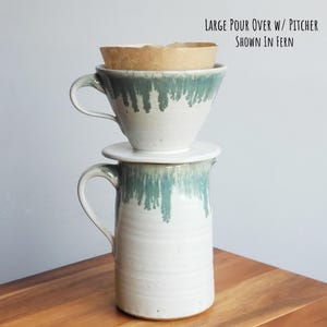May include: A white ceramic pour-over coffee maker and pitcher set with a teal glaze dripping down from the top. A brown paper filter sits inside the pour-over. The text "LARGE POUR OVER W/ PITCHER SHOWN IN FERN" is at the top.