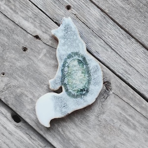 May include: A fox-shaped decorative object featuring a white and light gray speckled finish with a prominent, textured, dark green and crystalline center. The fox is depicted in a seated position with its tail curled forward along the side.