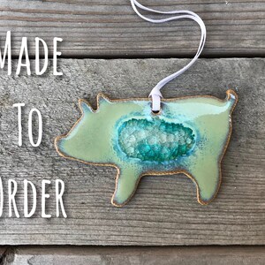 May include: A green ceramic pig ornament with a blue and white agate-like design in the center. The ornament has a white string attached for hanging. The text "MADE TO ORDER" is visible in the image.