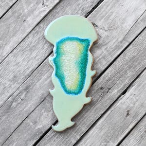 May include: A light green ceramic jellyfish with a blue and green center. The jellyfish is on a wooden background.