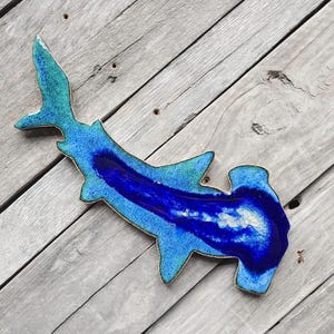 May include: A blue and green ceramic hammerhead shark wall decor. The shark is painted with a blue and green glaze, and the details are highlighted with gold paint.