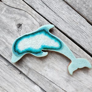 May include: A light blue ceramic dolphin figurine with a white and green swirl design. The dolphin is laying on a weathered wooden surface.