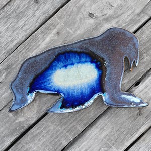 May include: A ceramic wall hanging in the shape of a walrus. The walrus is painted in shades of blue and black with a white accent.