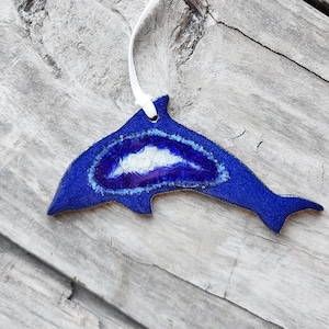 K&ouml;nnte beinhalten: Blaues, delfinf&ouml;rmiges Ornament mit wei&szlig;em Band zum Aufh&auml;ngen. Das Ornament hat eine blaue Au&szlig;enseite mit einem wei&szlig;en und lila Innendesign. Das Ornament liegt auf einer verwitterten Holzoberfl&auml;che.