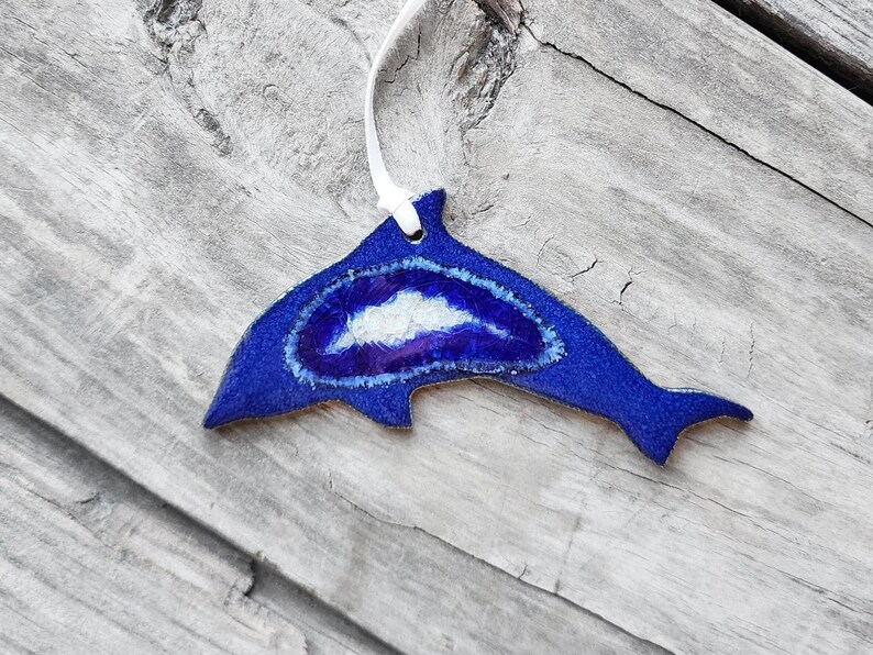 K&ouml;nnte beinhalten: Blaues, delfinf&ouml;rmiges Ornament mit wei&szlig;em Band zum Aufh&auml;ngen. Das Ornament hat eine blaue Au&szlig;enseite mit einem wei&szlig;en und lila Innendesign. Das Ornament liegt auf einer verwitterten Holzoberfl&auml;che.