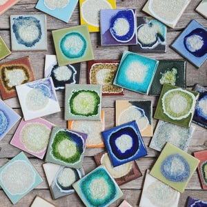 May include: A collection of square ceramic tiles featuring a central textured white circular glaze surrounded by vibrant colored borders in shades of blue, green, red, yellow, and purple, arranged in a scattered pattern on a wooden surface.