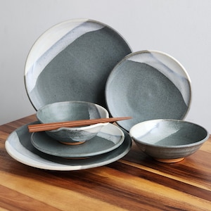 May include: A set of four ceramic bowls and plates in a gray and white glaze. The bowls and plates have a subtle, uneven texture and a white rim. The set includes a pair of chopsticks.