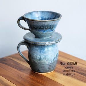 May include: A blue ceramic pour-over coffee set with a matching mug. The set includes a cone-shaped pour-over, a saucer, and a barrel-shaped mug. The text "SMALL POUR OVER" is visible in the image.