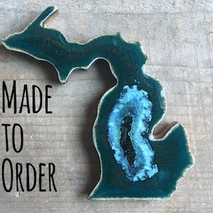May include: A teal blue ceramic Michigan state outline with a light blue center. The outline is on a wood background with the text "MADE TO ORDER" in black.