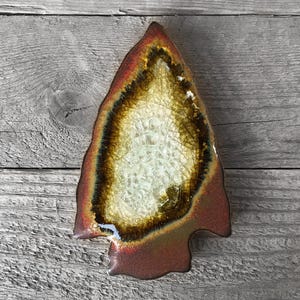 May include: A ceramic arrowhead shaped dish with a brown and white agate design. The dish has a red and orange glaze on the outside.