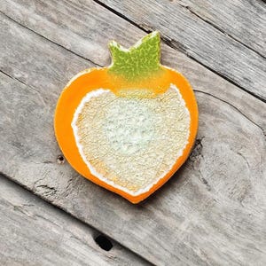 May include: A heart-shaped ceramic dish with an orange border and a textured, off-white center. The top has a green, textured stem. The dish is set on a weathered, gray wooden surface.