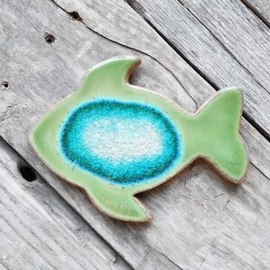 May include: A green ceramic fish with a blue and white center. The fish is on a wooden surface.