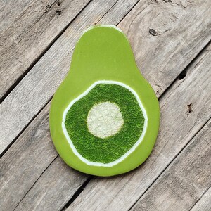 May include: A ceramic avocado-shaped coaster with a light green exterior and a white-bordered, textured green and white interior, set against a rustic wooden surface. The coaster is a decorative kitchen item.