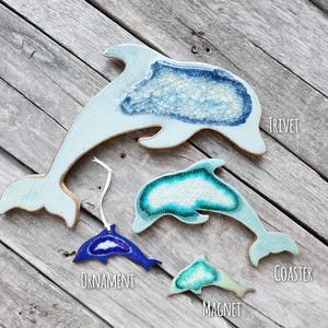K&ouml;nnte beinhalten: Eine Sammlung von Keramikartikeln in Delfinform, darunter ein Topfuntersetzer, ein Ornament, ein Untersetzer und ein Magnet. Die Artikel sind hellblau und wei&szlig; mit einem strukturierten, meerartigen Design. Das Ornament ist dunkelblau.
