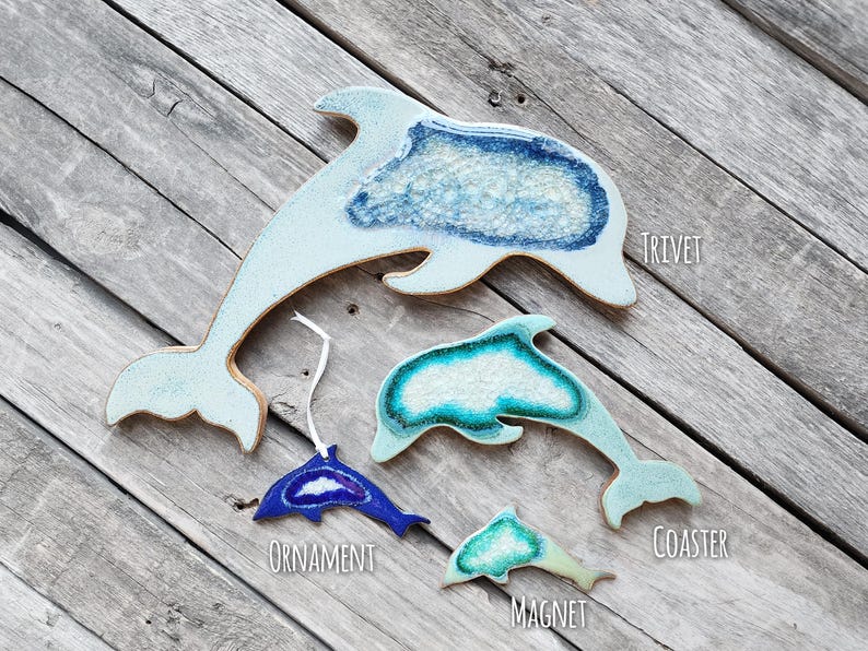K&ouml;nnte beinhalten: Eine Sammlung von Keramikartikeln in Delfinform, darunter ein Topfuntersetzer, ein Ornament, ein Untersetzer und ein Magnet. Die Artikel sind hellblau und wei&szlig; mit einem strukturierten, meerartigen Design. Das Ornament ist dunkelblau.