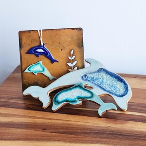 K&ouml;nnte beinhalten: Dekorative Keramik-Delfin-Kunst in Blau- und Wei&szlig;t&ouml;nen. Das Kunstwerk umfasst einen gro&szlig;en Delfin und zwei kleinere, mit einer strukturierten, glasierten Oberfl&auml;che. Die Delfine werden mit einem braunen Hintergrund pr&auml;sentiert.