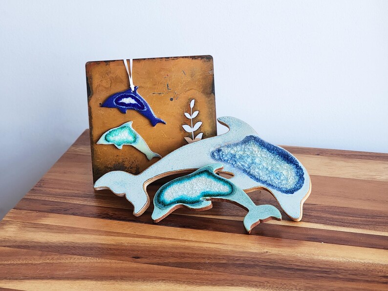 K&ouml;nnte beinhalten: Dekorative Keramik-Delfin-Kunst in Blau- und Wei&szlig;t&ouml;nen. Das Kunstwerk umfasst einen gro&szlig;en Delfin und zwei kleinere, mit einer strukturierten, glasierten Oberfl&auml;che. Die Delfine werden mit einem braunen Hintergrund pr&auml;sentiert.