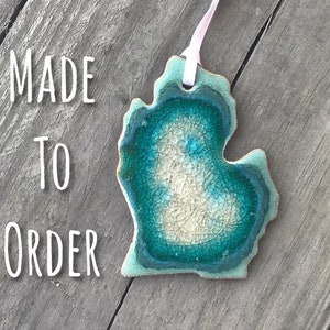 May include: A ceramic ornament shaped like the state of Michigan, with a blue and green glaze. The ornament has a white ribbon attached to it and the text "MADE TO ORDER" is visible in the image.