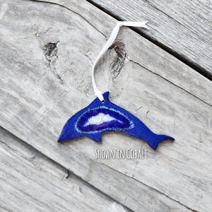 K&ouml;nnte beinhalten: Ein kobaltblaues, delfinf&ouml;rmiges Ornament mit einem wei&szlig;en Band zum Aufh&auml;ngen. Der Delfin hat ein zentrales Design mit wei&szlig;en und violetten Akzenten. Der Text "SHOWN IN COBALT" ist sichtbar.