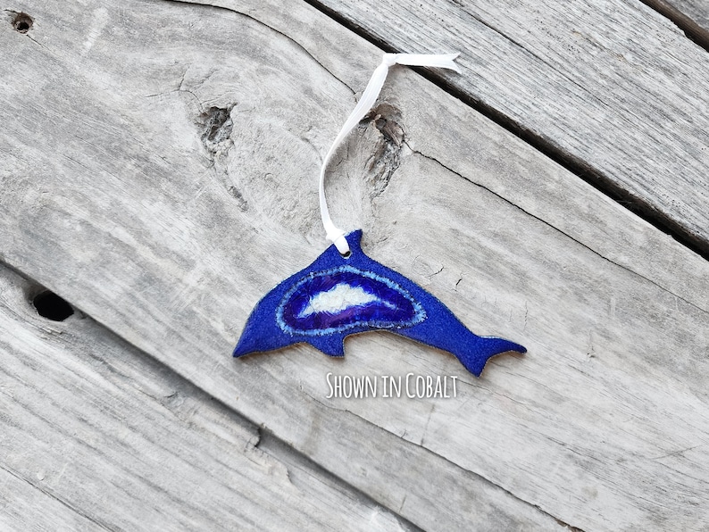 K&ouml;nnte beinhalten: Ein kobaltblaues, delfinf&ouml;rmiges Ornament mit einem wei&szlig;en Band zum Aufh&auml;ngen. Der Delfin hat ein zentrales Design mit wei&szlig;en und violetten Akzenten. Der Text "SHOWN IN COBALT" ist sichtbar.