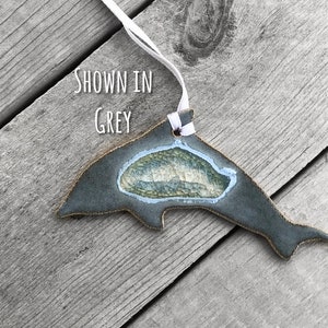 K&ouml;nnte beinhalten: Graues Keramik-Delphin-Ornament mit wei&szlig;em Band zum Aufh&auml;ngen. Der Delphin hat eine strukturierte, hellblaue und gr&uuml;ne Mitte. Der Text "SHOWN IN GREY" ist sichtbar. Das Ornament liegt auf einer verwitterten Holzoberfl&auml;che.