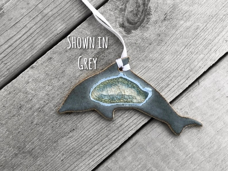 K&ouml;nnte beinhalten: Graues Keramik-Delphin-Ornament mit wei&szlig;em Band zum Aufh&auml;ngen. Der Delphin hat eine strukturierte, hellblaue und gr&uuml;ne Mitte. Der Text "SHOWN IN GREY" ist sichtbar. Das Ornament liegt auf einer verwitterten Holzoberfl&auml;che.