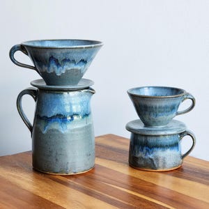 May include: A ceramic coffee set in shades of blue and gray. The set includes a pitcher, two cups, and two saucers. The pitcher is approximately 7 inches tall, and the cups are about 3 inches tall. The set has a glossy finish and a dripped glaze effect.