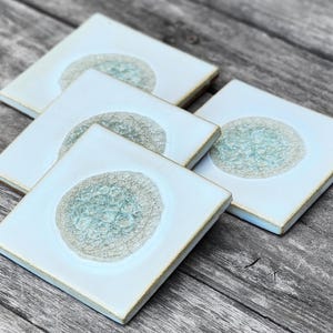 May include: Four white ceramic coasters with a light blue, textured, circular design in the center of each coaster.