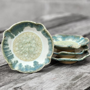 May include: Four small, scalloped ceramic dishes in shades of blue, green, and white. The dishes are stacked on top of each other, with the top dish featuring a textured, white center.