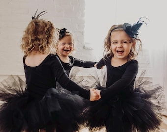 May include: Three children wearing black long-sleeved leotards with voluminous black tulle skirts. Each child wears a small black hat accessory with feathers on a headband. The children wear striped socks in orange and black or purple and black patterns.
