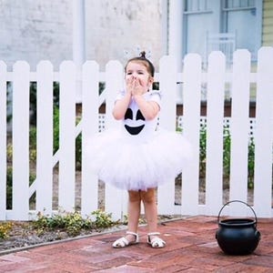 May include: A white ghost-themed costume featuring a short-sleeved top with a black ghost face graphic and a layered white tulle tutu skirt, paired with white sandals and a small black plastic cauldron bucket for collecting treats.