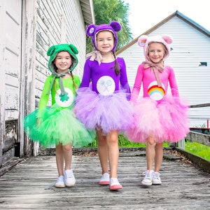 May include: Three girls dressed as Care Bears for Halloween. The girl on the left is wearing a green tutu and a green shirt with a shamrock on it. The girl in the middle is wearing a purple tutu and a purple shirt with a lollipop on it. The girl on the right is wearing a pink tutu and a pink shirt with a rainbow on it.