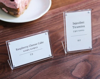 May include: Two white acrylic sign holders with black text on a wooden surface. The sign on the left reads "Raspberry Cheese Cake" with the Korean translation and "7.0". The sign on the right reads "Injeolmi Tiramisu" with the Korean translation and "8.0".