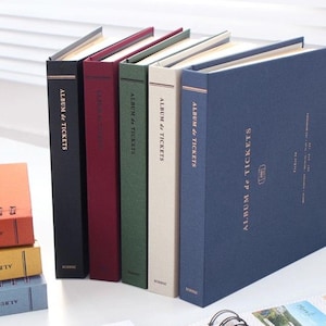 May include: A selection of ticket albums in colours such as black, burgundy, green, cream, and navy blue. The albums are arranged vertically, with the text "ALBUM de TICKETS" on the spine. Additional albums are stacked in the foreground.