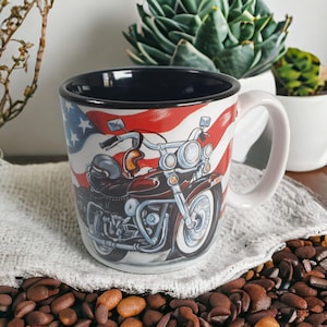 May include: White ceramic mug featuring a motorcycle design against a backdrop of the American flag. The mug has a black interior and a white handle. The motorcycle is detailed with chrome accents and a helmet.