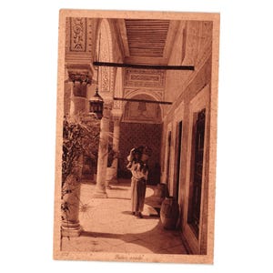 May include: A sepia-toned vintage postcard depicting a courtyard with stone columns, a hanging lantern, and a person carrying a vessel on their head. The architecture features intricate wall carvings and tiled patterns. The text reads Palais arabe.