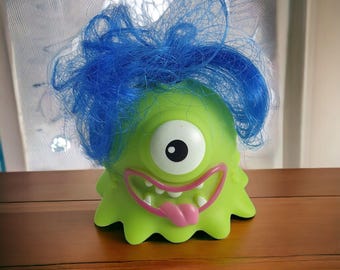 May include: A lime green monster toy with a single large eye, a wide grin, and a pink tongue. The monster has blue, messy hair. The toy is sitting on a wooden surface.