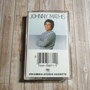 May include: A retro cassette tape of Johnny Mathis. The cover displays a smiling man in a grey sweater. The clear plastic case has the text "JOHNNY MATHIS" and "COLUMBIA STEREO CASSETTE".