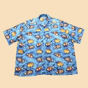 Puede incluir: Camisa de manga corta con fondo azul y estampado de peces amarillos, naranjas y marrones. La camisa tiene cuello clásico y corte relajado. El estampado de peces se repite por toda la tela.