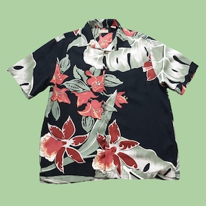 Puede incluir: Camisa negra de manga corta con estampado floral. La camisa presenta grandes flores rojas y blancas y hojas verdes. Tiene una botonadura frontal y un cuello. La camisa está hecha de una tela ligera.