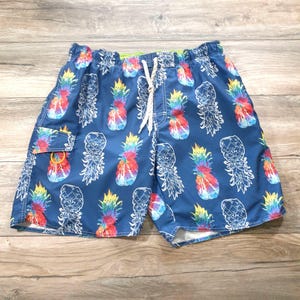 Könnte beinhalten: Blaue Badeshorts mit einem farbenfrohen Ananasmuster. Die Shorts haben einen Kordelzug in der Taille und eine kleine Seitentasche. Die Ananas sind in Rot-, Gelb-, Blau- und Grüntönen mit weißen Umrissen.
