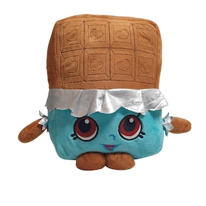 May include: A plush toy shaped like a chocolate bar with a smiling face. The top section is brown with heart-shaped details, while the bottom is blue with large red eyes and a silver foil wrapper.