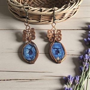 May include: A pair of handcrafted copper wire earrings. Each earring features a blue stone with a dark blue design, encased in a copper wire frame. The earrings have a decorative swirl design at the top and a hook closure. A wicker basket and lavender sprigs are in the background.
