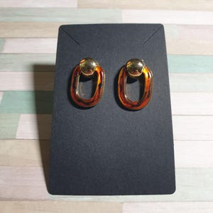 May include: A pair of oval-shaped earrings with a tortoiseshell pattern and gold-colored accents. The earrings are displayed on a black card against a light-colored background.