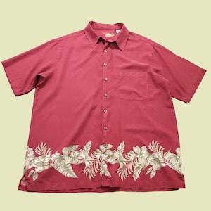 Pode incluir: Camisa de manga curta com botões em tom vermelho escuro. A camisa apresenta um padrão floral ao longo da borda inferior, com folhas e flores brancas e verdes. Possui um bolso no peito e uma gola clássica.