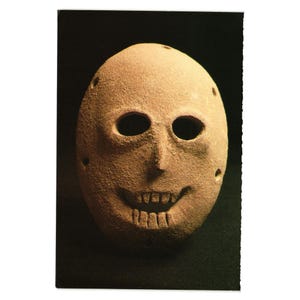 May include: A tan-colored, oval-shaped mask with large, dark eye holes and a smiling mouth. The mask has several small holes around the edges and a textured surface, suggesting it is made of stone or a similar material. The background is dark.