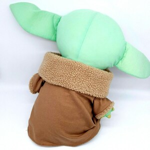 Puede incluir: Un peluche de un personaje con grandes orejas verdes y una cabeza verde. El personaje viste un atuendo marr&oacute;n con un cuello beige y difuso. El juguete est&aacute; dise&ntilde;ado para parecerse a un personaje popular de una serie de ciencia ficci&oacute;n.