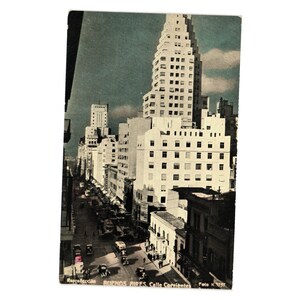 May include: A vintage postcard featuring a high-angle view of Calle Corrientes in Buenos Aires. The scene shows white multi-story buildings, including a tall skyscraper, lining a busy street with vintage cars and pedestrians. Text reads: BUENOS AIRES. Calle Corrientes.