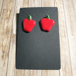 May include: A pair of red pepper-shaped earrings with green stems. The earrings are displayed on a black card against a light wood grain background. The earrings are a bright red color.