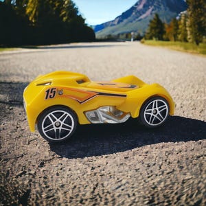 May include: A bright yellow toy race car with the number "15" on the side. The car has black and silver wheels and orange and black detailing. The car is sitting on a paved road with a mountain in the background.